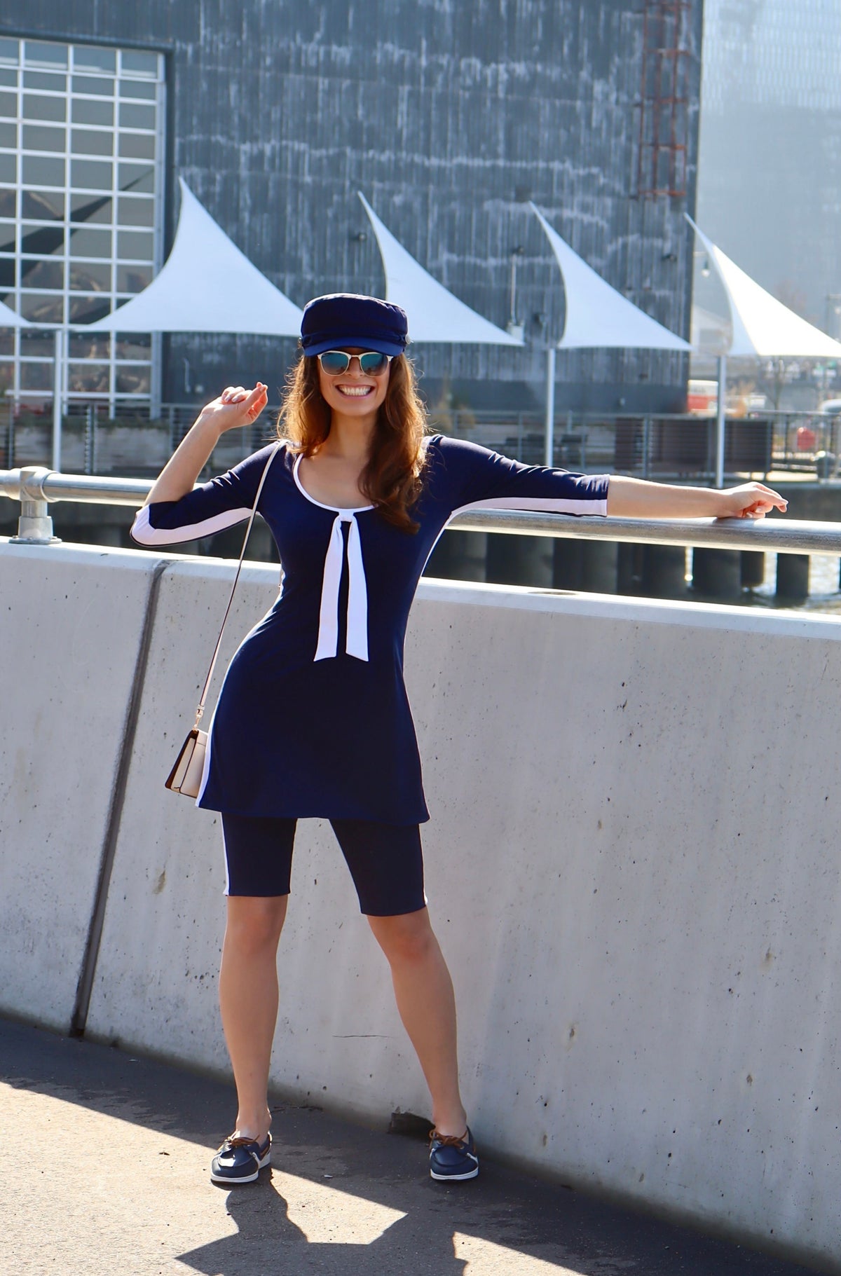 Model wearing navy with white stripes down the sides, 3/4 sleeves and a white tie tunic and a blue hat with one hand on a railing smiling.