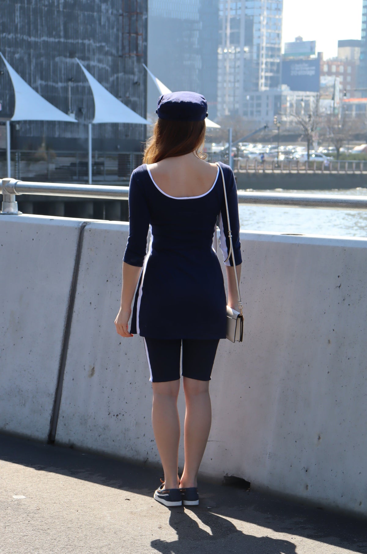 Back of model wearing navy with white stripes down the sides, 3/4 sleeves and a white tie tunic and a blue hat.