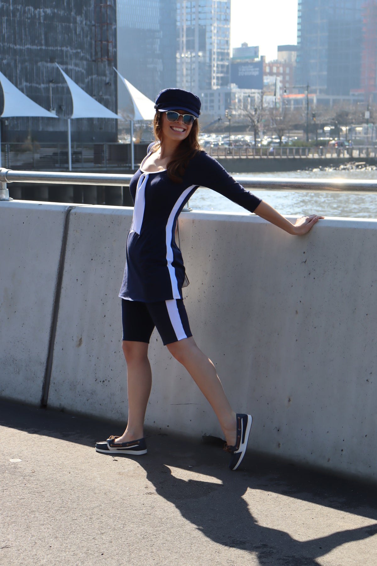 Model wearing navy bike shorts with white stripes down the sides, navy with white stripes down the sides, 3/4 sleeves and a white tie tunic and a blue hat smiling in front of the water, reaching back.