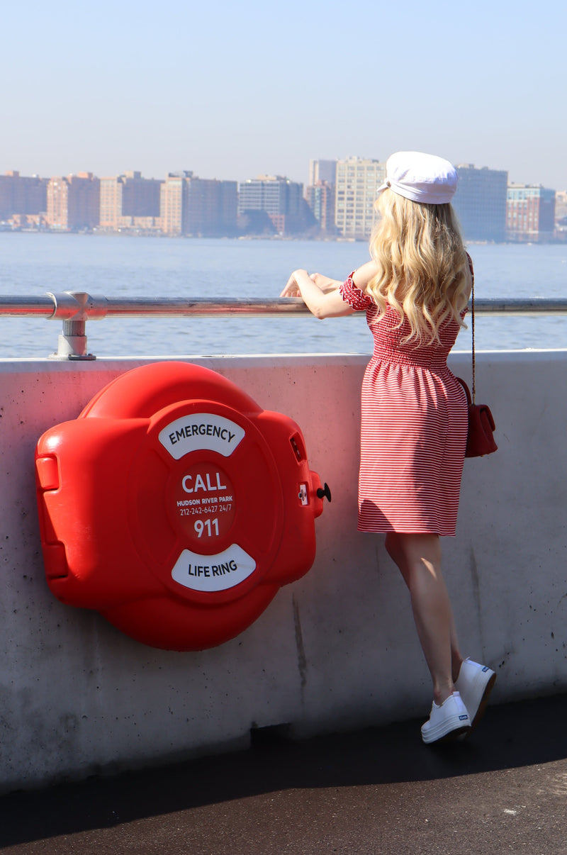 Back of model wearing red and white striped shirred dress and white hat looking out towards the water.