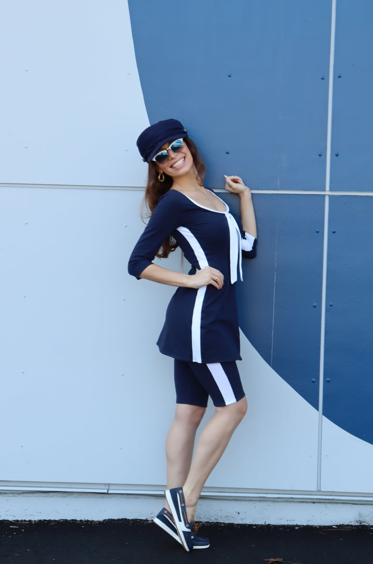 Model wearing navy with white stripes down the sides, 3/4 sleeves and a white tie tunic and a blue hat in front of a blue wall with one hand on her hip smiling