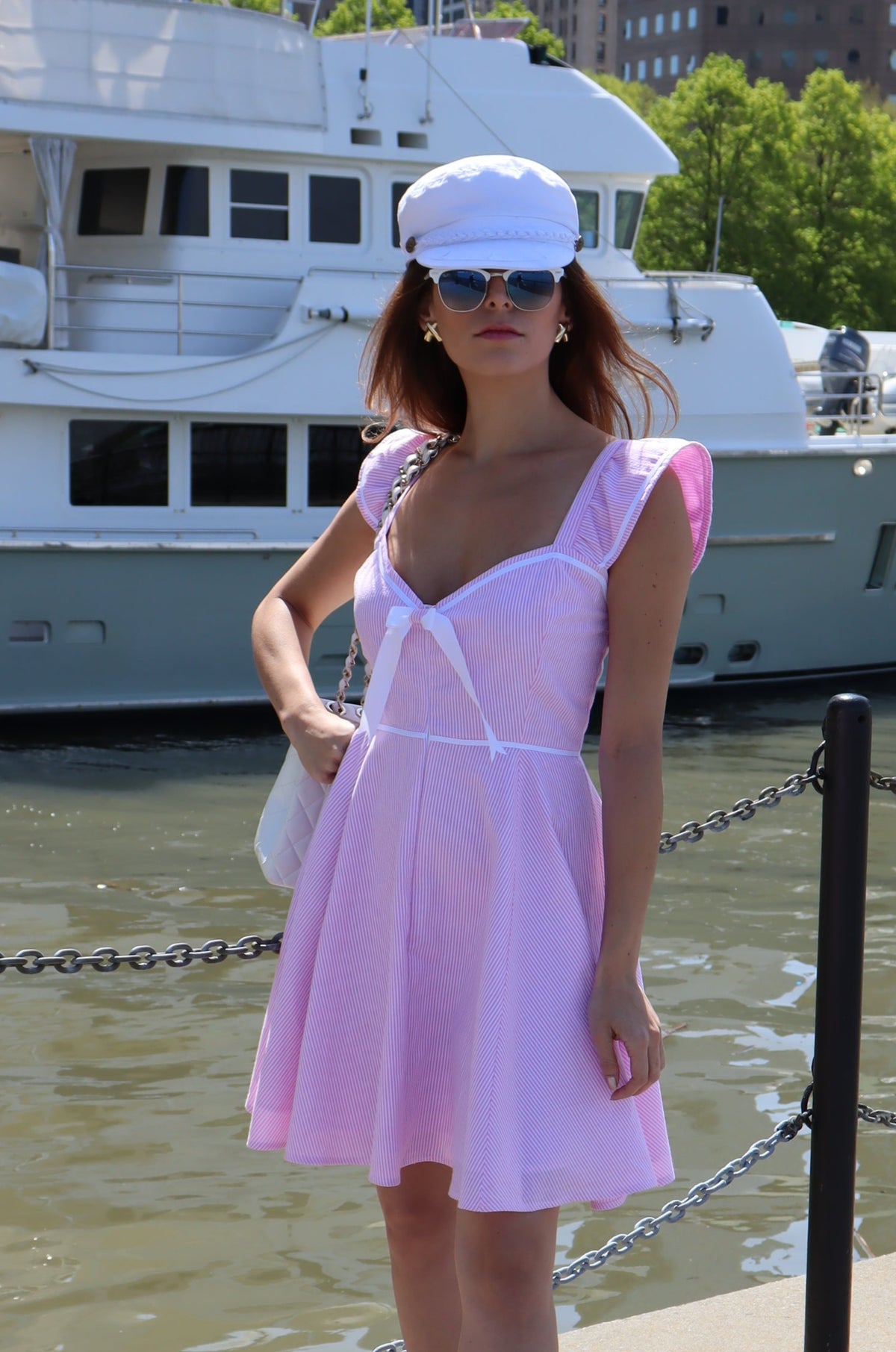 Model wearing a white hat and a pink and white seersucker dress with a white bow in front of a white boat.