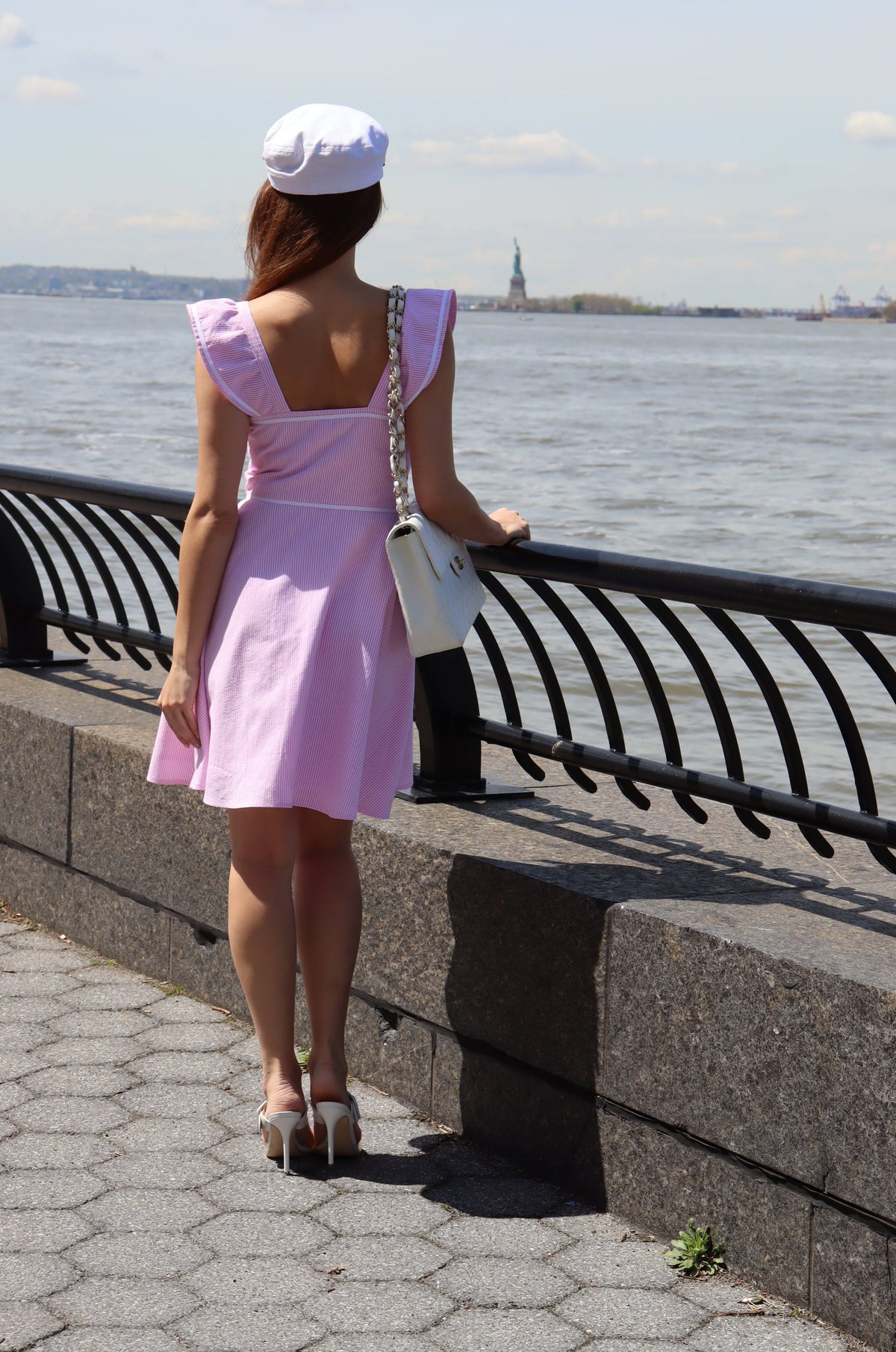 Back of model wearing a white hat and a pink and white seersucker dress with a white bow in front of water.