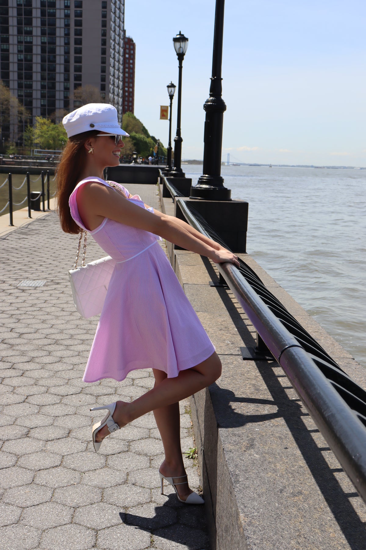 Model wearing a white hat and a pink and white seersucker dress with a white bow looking out to the water.