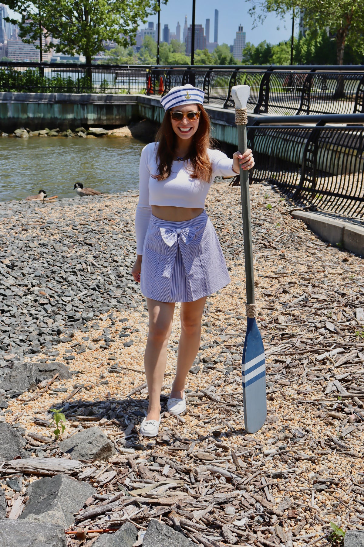 Model wearing a white and blue striped sailing hat, a white long sleeve crop top, and a light blue cotton print skirt with a bow, holding an oar smiling.