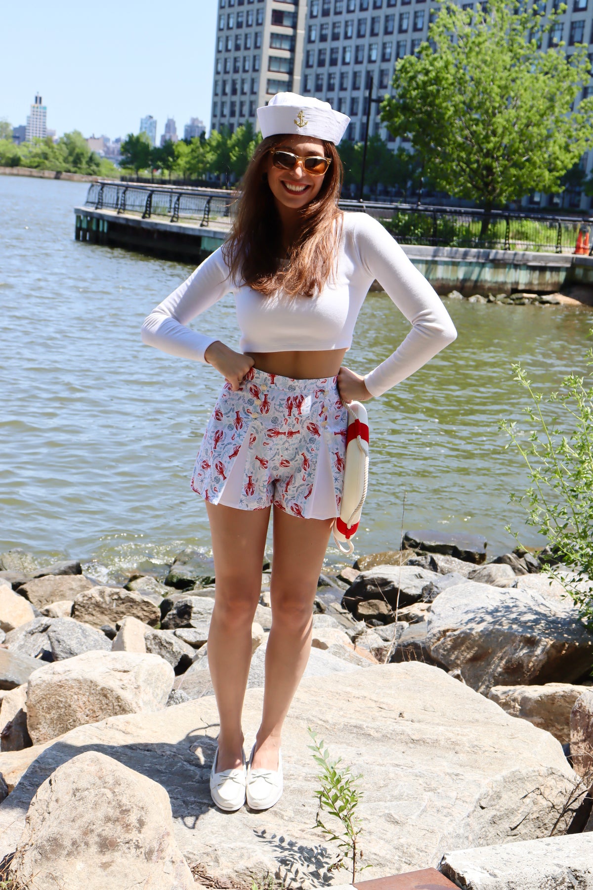 Model wearing pleated blue and red lobster print cotton shorts and white long sleeve crop top with her hands on her hips, standing on rocks in front of a river, smiling.