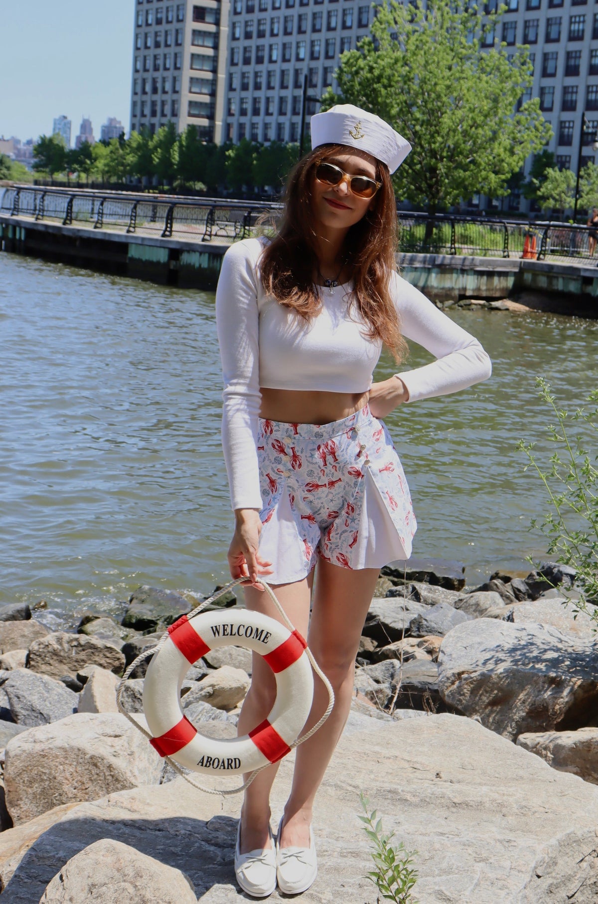 Model wearing pleated blue and red lobster print cotton shorts and white long sleeve crop top, with her hand on her hip, standing on rocks in front of a river, holding a a red and white buoy.
