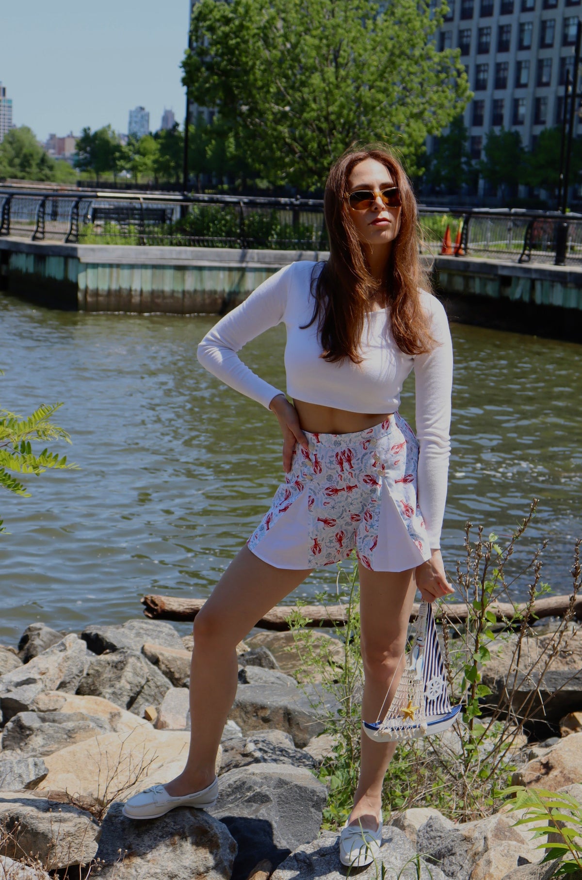 Model wearing pleated blue and red lobster print cotton shorts and white long sleeve crop top with her hand on her hip looking off to the side, standing on rocks in front of a river.