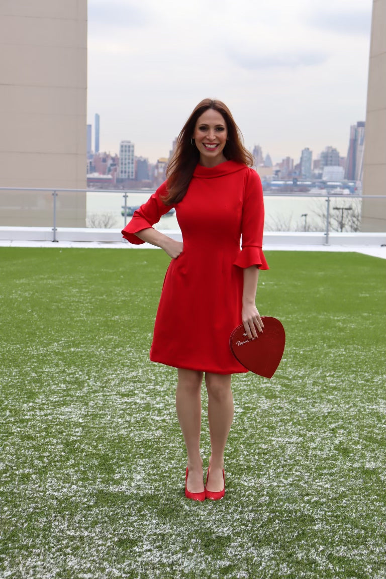 Model wearing a red dress with 3/4 sleeves, with a hand on her hip and holding a heart shaped box smiling.