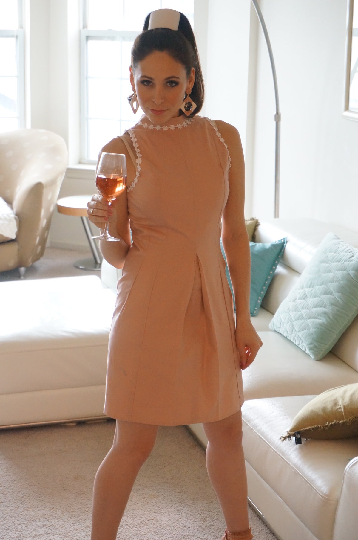  Model is wearing a pink dress with lace daisy trim and a front kick pleat looking off to the side in front of a couch holding a wineglass.